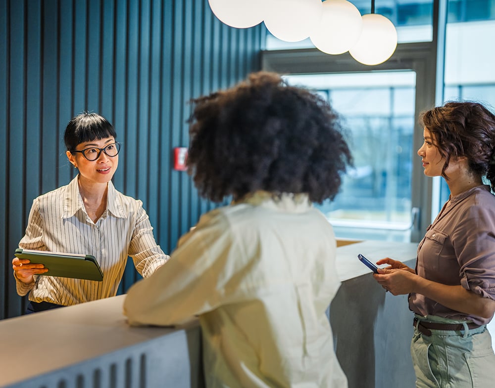 A hotel front desk scene where a woman clerk stands behind the counter helping two female customers.