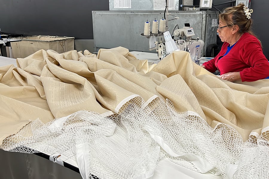 Worker sewing a beige privacy curtain on an industrial sewing machine.