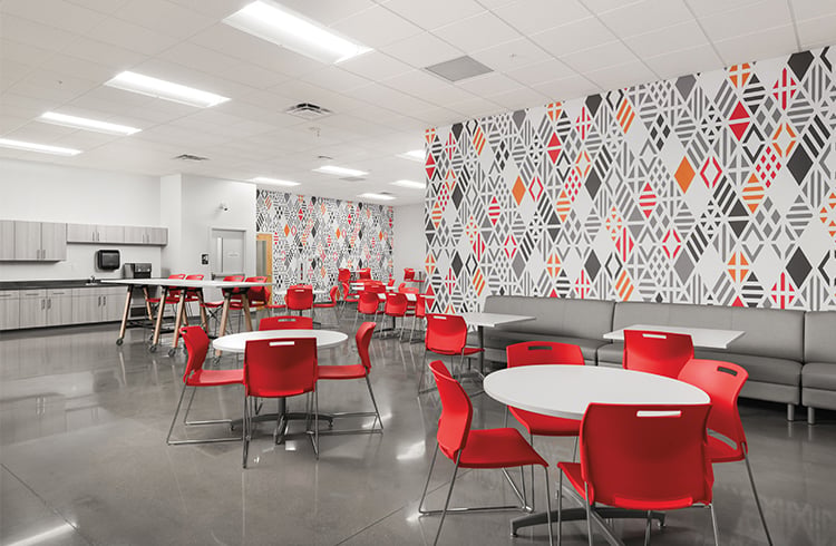 Spacious school common area with glossy floors, round tables surrounded by red chairs, gray booth seating, and a large wall decorated with geometric patterns in gray, orange, and red.