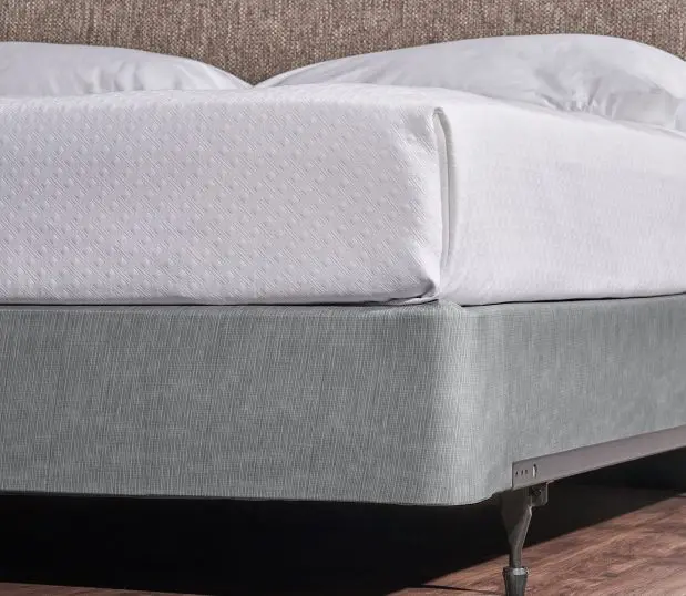 Encircle Box Spring Cover in the color Magnesium. Shown on a king size bed with a white top cover and tan head board with wood flooring and metal bed frame.