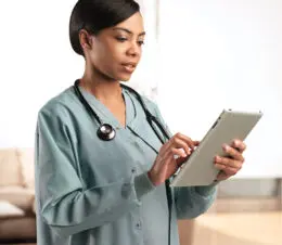 A doctor looking at an iPad is shown wearing an Excel® Unisex Warm Up Jacket shown in Ceil.