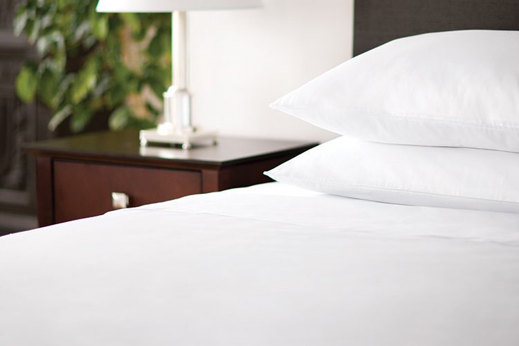 A hotel bed next to nightstand with Centium Satin sheets and two pillows.