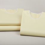 A folded ComPel Precaution gown leaning agains a stack of two additional folded gowns.