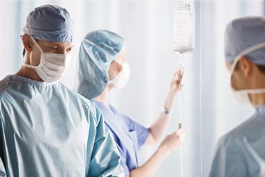Three surgical team members stand in an operating room. They are wearing surgical gowns and face masks. One is seen adjusting an IV.