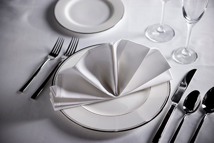 A well-set dining table, a ornately folded napkin sits on the plate.