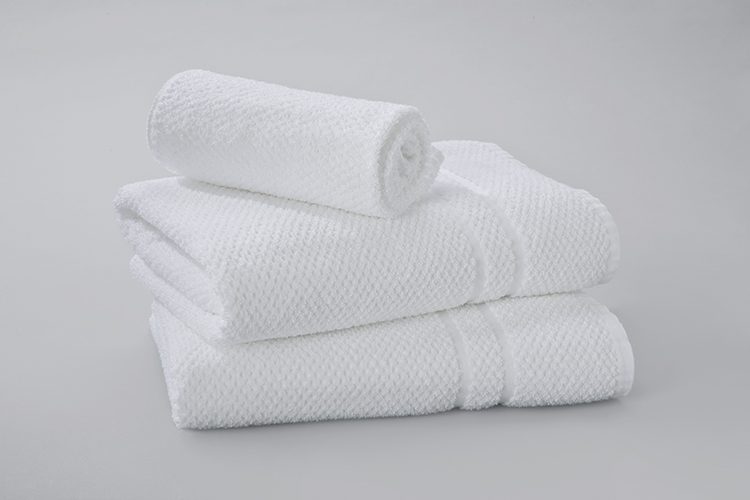 A rolled Pique Weave wash towel rests atop a stack of two folded Pique Weave bath towels.