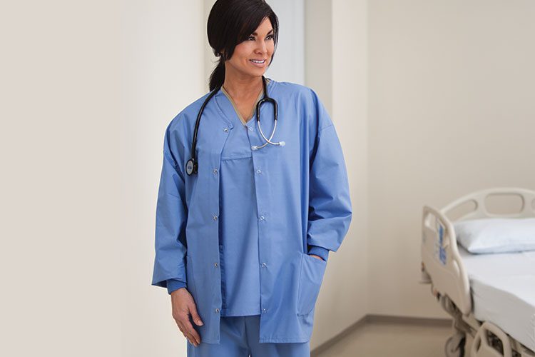 A woman standing in a patient room, wearing blue hospital scrubs and a a warm-up jacket. These medical scrubs (or medical apparel) are made of Softweave®. We also offer unisex scrubs tops and pants.