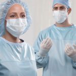 Two doctors prepping for surgery, wearing surgical gowns, masks, gloves, and hair protection.