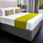 A hotel guest bed made with a custom bed scarf and two custom made decorative throws pillows.