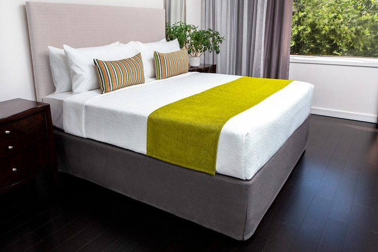 A hotel guest bed made with a custom bed scarf and two custom made decorative throws pillows.