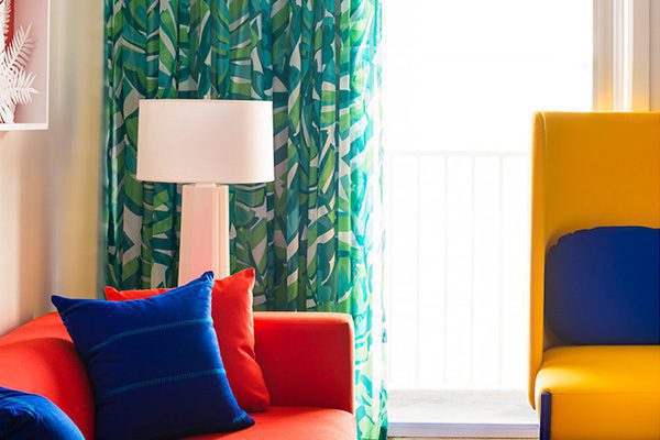 A vibrant seating area in a hotel room, featuring a red sofa, yellow chair, and green window curtains.