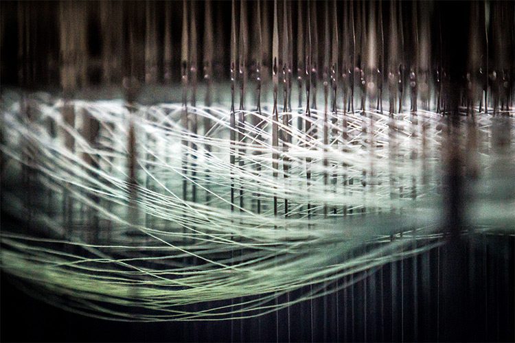 A detailed image of a textile weaving machine, dozens of threads can be seen flowing from left to right.
