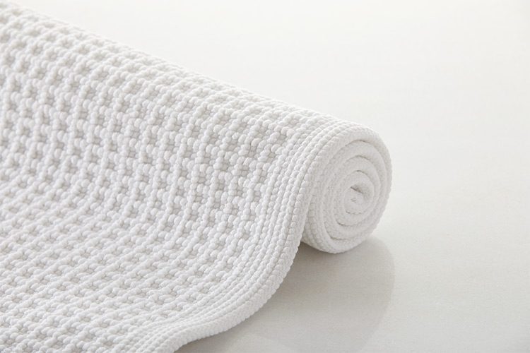A white, textured bath rug, partially rolled up, lays on a white background.