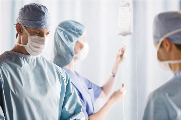 Three surgical team members stand in an operating room. They are wearing reusable surgical gowns. One is seen adjusting an IV.