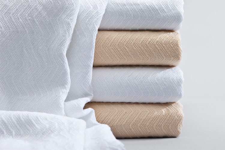 A white Herringbone wholesale hotel blanket is draped over a folded stack of Herringbone blankets for hotels.