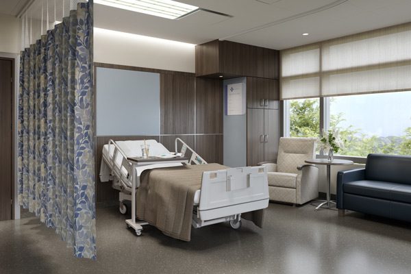 A patient room in a hospital with hospital bed, privacy curtains in a blue pattern and layered blinds for privacy and light control..