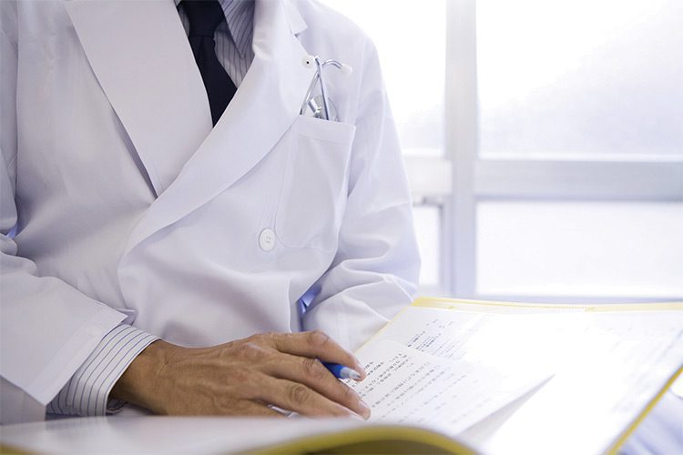 A close up image of a doctor wearing a lab coat, making notes on a patient chart. Unisex, men's & women's lab coats are sold by Standard Textile.