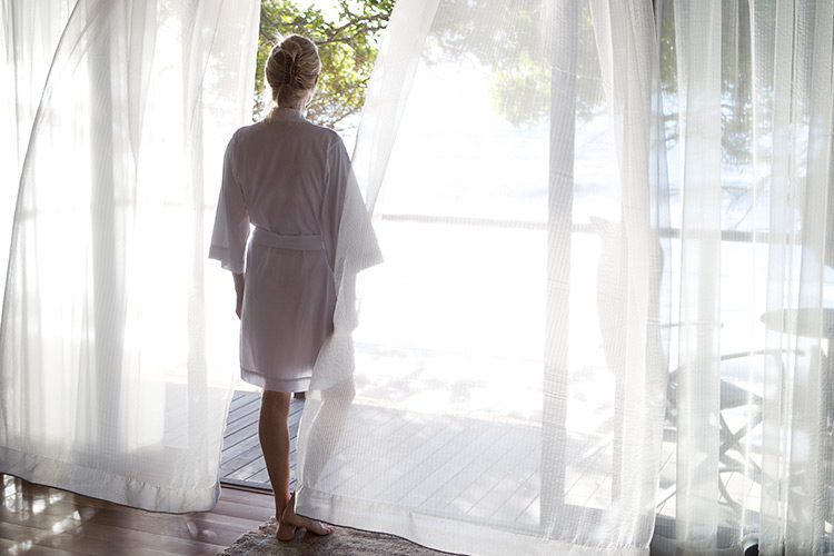 A woman in a bath robe stands, staring out a large floor to ceiling glass patio door.