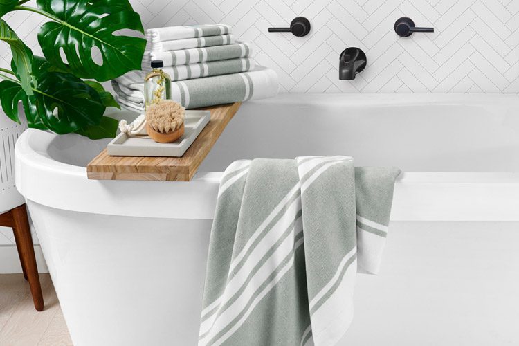 A large white bath tub, a bath towel draped over the side, and a stack of folded bath towels, and hand towels stacked nearby.