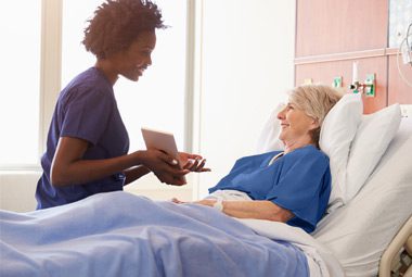 Nurse conversing with a patient in hospital bed. The bed has DermaTherapy® sheets.