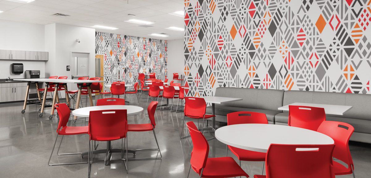 Image of a cafeteria with white tables, bright red chairs and bold wallcovering.