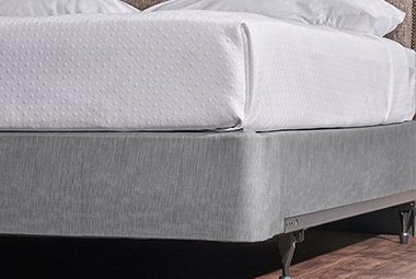 Encircle Box Spring Cover in Magnesium shown on a wooden floor with a metal bed frame with white top cover.