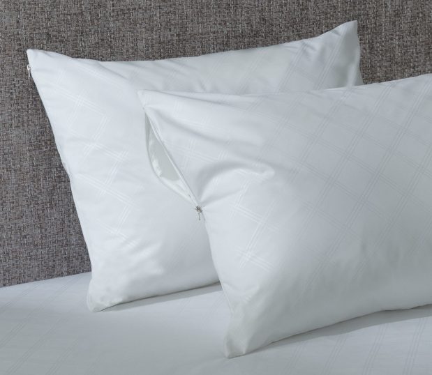 Two pillows featuring antimicrobial pillow covers are stacked against a headboard on a bed.