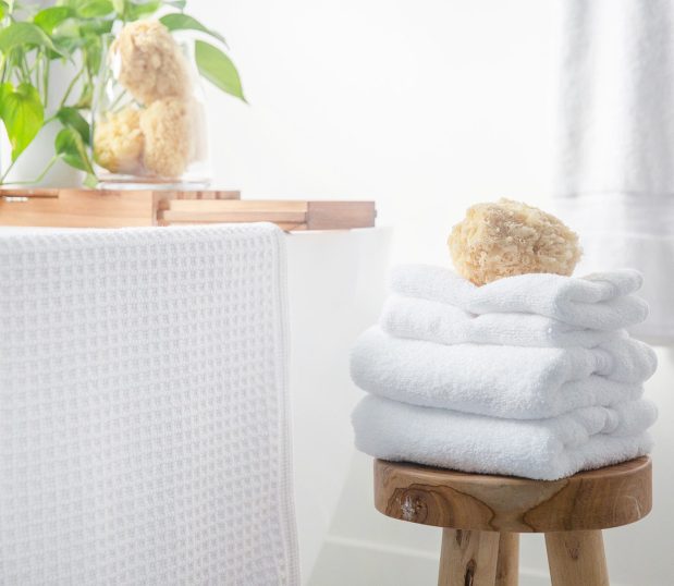 This cotton bath rug adds luxury to any bathroom. We use only 100% high-quality cotton, bold textures, and tufted fabric for a plush experience. An Artesano bath rug is shown draped on a tub next to a stack of plush Lynova towels.