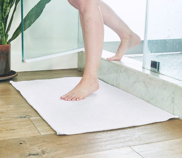 This cotton bath rug adds luxury to any bathroom. We use only 100% high-quality cotton, bold textures, and tufted fabric for a plush experience. Image shows a person stepping out of the shower onto an Artesano bath rug.