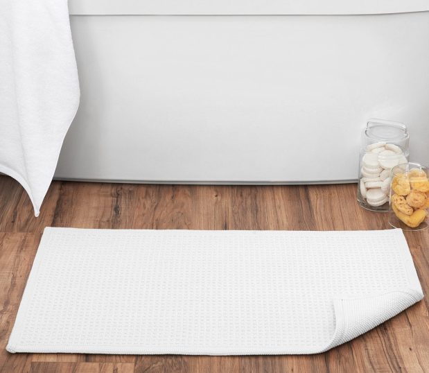 Image shows an absorbent Artesano bath rug on the floor in front of a bath tub.