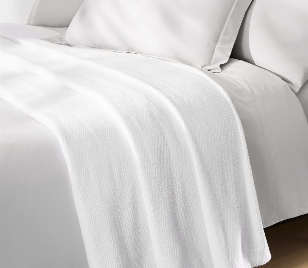 This all-purpose bed blanket is perfect for all seasons and climates. Luxurious 100% cotton provides hotel guests with warm comfort and breathability. Shown here: a white blanket on a bed.