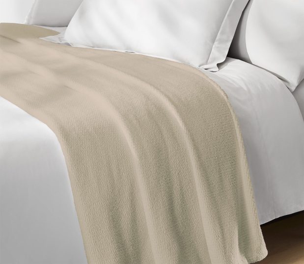 This all-purpose bed blanket is perfect for all seasons and climates. Luxurious 100% cotton provides hotel guests with warm comfort and breathability. Shown here: a champagne Lynova blanket laying on a bed.