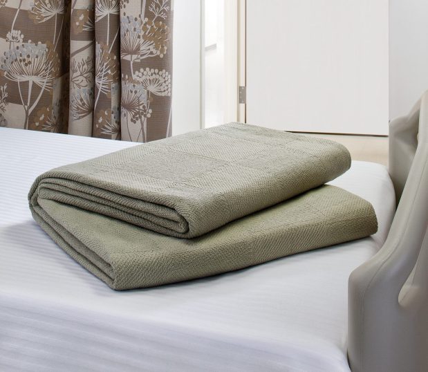 A Serenity™ Spread blanket is shown laying folded on a hospital bed. With large woven squares this traditional hospital blanket is shown here in Olive.