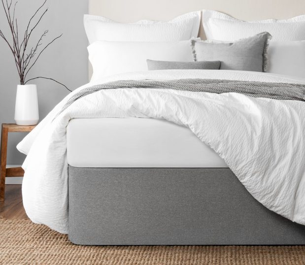 This is an image of the Circa® Bed Wrap on a beautifully made bed. The bed has a fluffy comforter, white sheets and a gray knit throw on a boutique hotel bed.