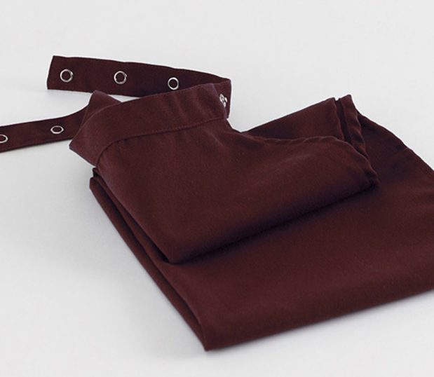 A burgundy Dignity Clothing Protector is neatly folded in this image. This adult clothing protector has multiple snaps at the neck to easily adjust for the correct fit.