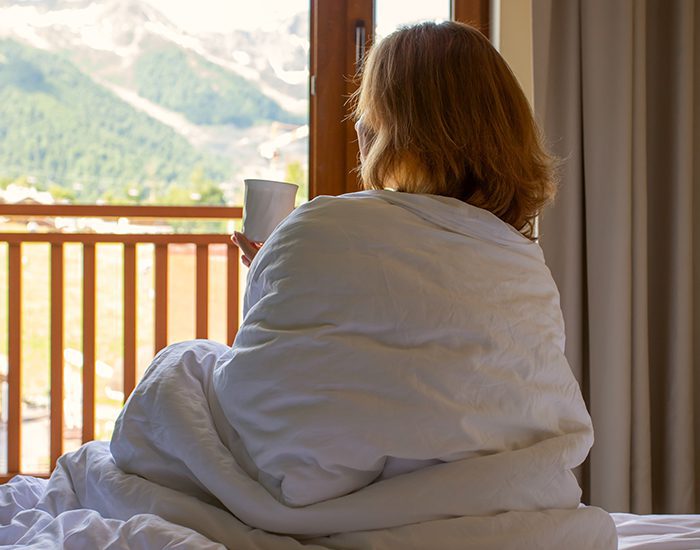 A woman wrapped in a duvet and sipping coffee sits on a white hotel bed and enjoys the scenery outside the window.