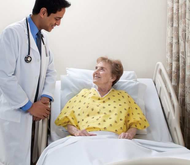 Doctor speaking with female patient wearing a Risk Identity Patient Gown. This hospital gown is a lapover V-neck style made from Champion Cloth fabric, with I.V. sleeves and telemetry pockets in Kaleidoscope Yellow color.