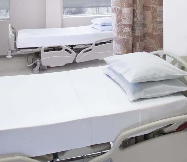 TruVal® Hospital Sheets for hospitals and health systems. Shown here in a hospital room on two hospital beds with contour and flat sheets, and two stacked pillows in pillowcases.