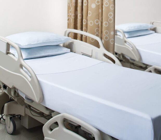 UniVal® Woven Hospital Sheets no-iron, crease-resistant sheets for hospitals and health systems. Shown here in a hospital room on two hospital beds with contour and flat sheets, and two stacked pillows in pillowcases.