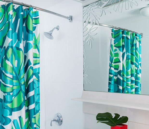 Custom shower curtain in a tropical pattern in blue, green and teal is shown hanigng from a shower rod in a hotel bathroom.