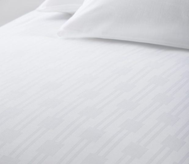 Image of the White Reed Top Cover on a bed with a couple of white pillows. This top sheet has a subtle stripe and square design. A top cover is more easily laundered than a bedspread and will make for a more sanitary and hygienic room.