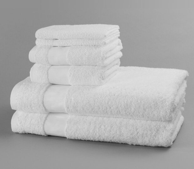 Two each of white absorbent bath towels, hand towels and washcloths stacked and shot against a grey backgrouond.