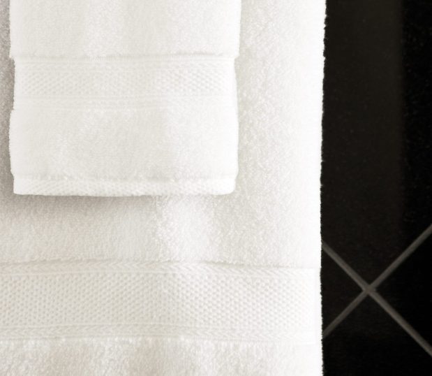 A white Lynova® microcotton towel and hand towel are hanging from a towel rod in a bathroom with a black tile wall.