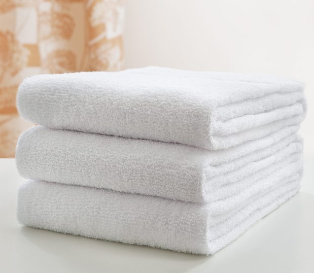 A stack of three folded PerVal® bath towels is shown on a hospital bed with a cubicle curtain in the background.