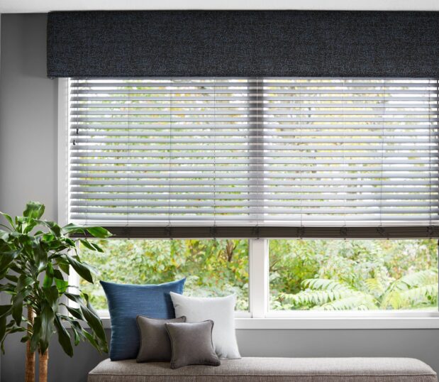 Custom cornices and top treatments such as valences, roman shades, and swags will create a unique room. This rooms window has a gray valance and window shade. Below the window is an ottoman with throw pillows.