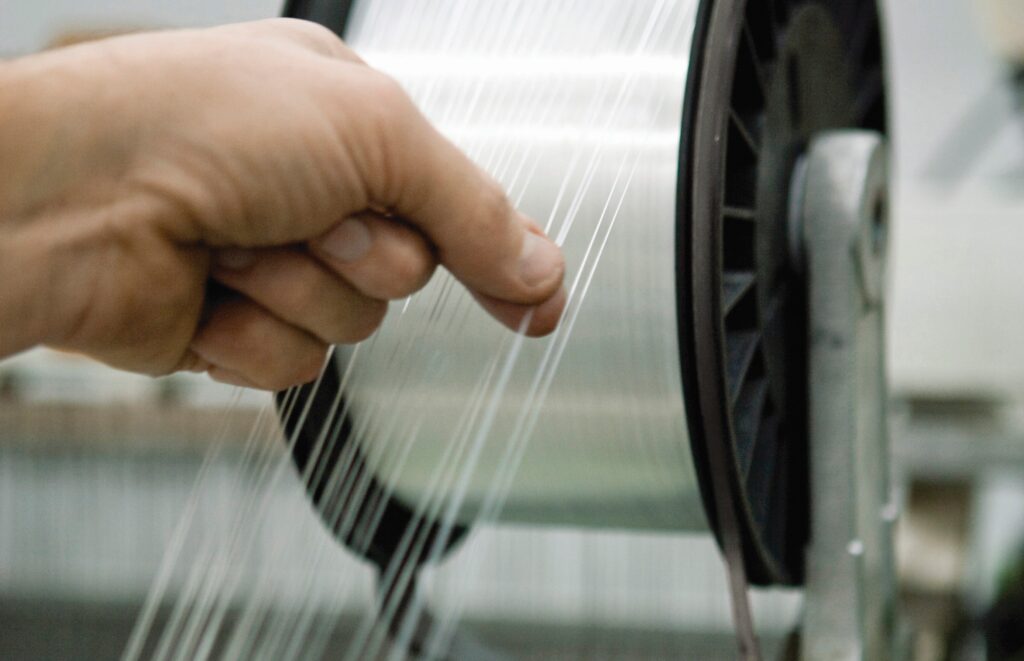A hand pinches several threads of 100% cotton yarn as they unwind from a spool.