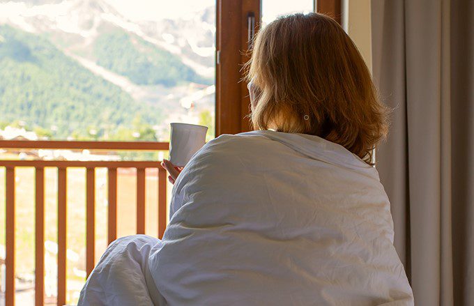 Woman sitting on a bed with coffee looking out at mountain scenery.