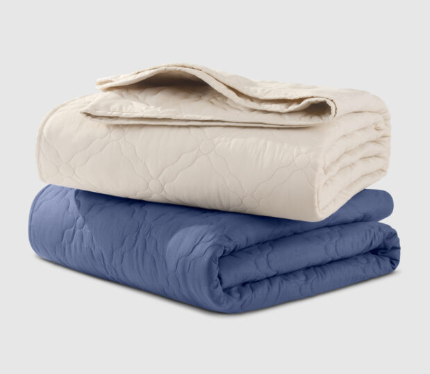 A stack of the Chateau latticed quilted bedspreads are shown here in both colorways Birch and Twilight.